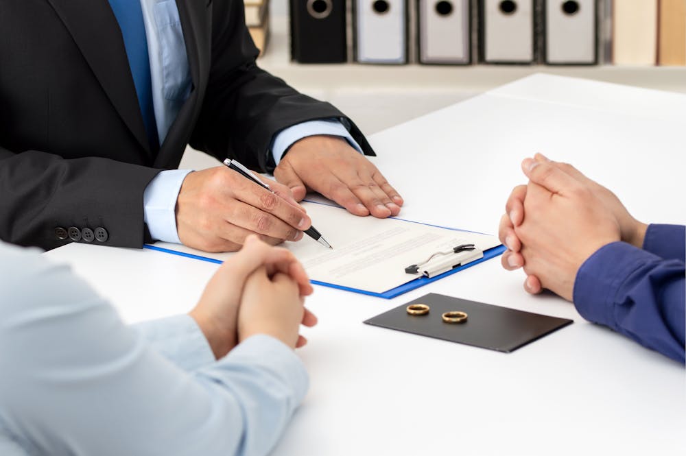 A man in a suit writes on a clipboard, while two individuals sit with clasped hands at a table. Two wedding rings rest on a black folder nearby, suggesting a divorce discussion.