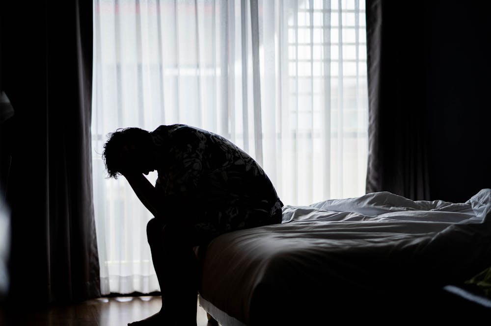 A person sits on a bed, head down and hands covering their face, silhouetted by light filtering through sheer curtains. The room appears dim and somber.
