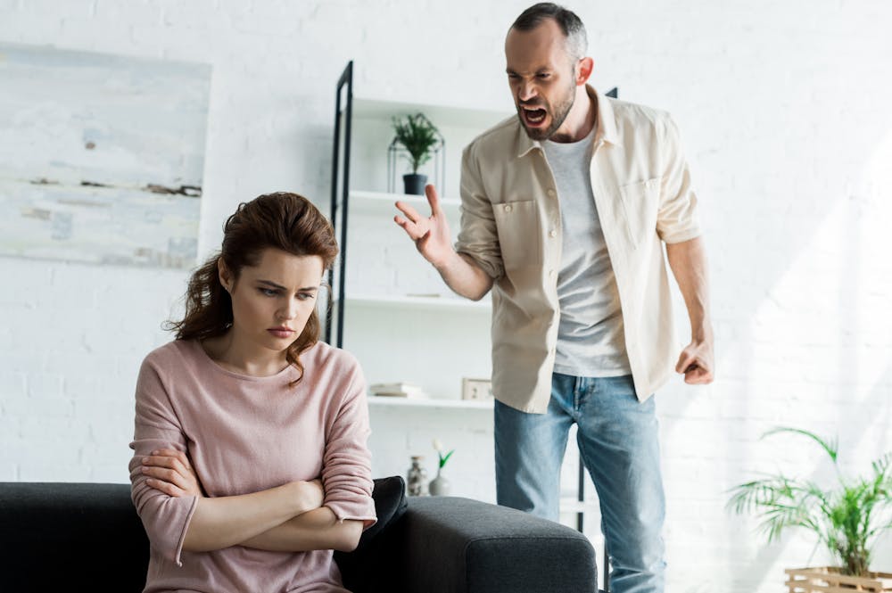 A woman sits with her arms crossed and a downcast expression on a couch, while a man stands nearby, yelling and gesturing angrily. The background features shelves with plants and decor.