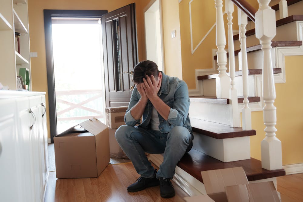 Man sitting with head in hands on staircase, surrounded by unpacked boxes, in a hallway with a yellow painted wall and an open door leading outside.