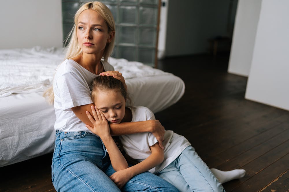 Woman sitting on wooden floor embraces a young girl, who leans her head on the woman's lap. Both wear white shirts and jeans. They are beside a white bed in a dimly lit room.