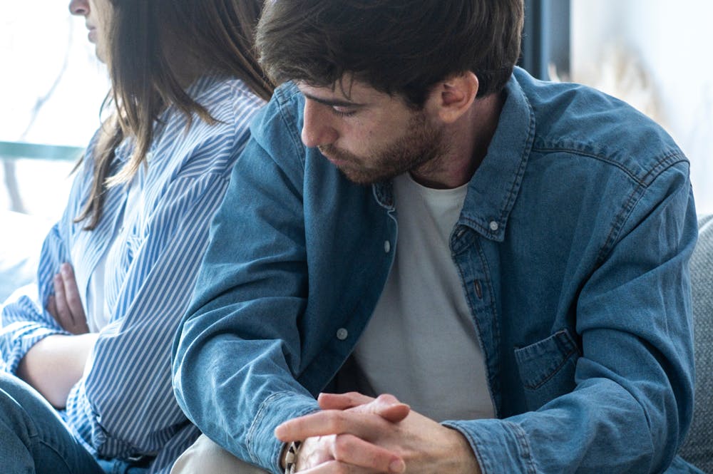 A man in a denim shirt sits with folded hands, looking down. A woman in a striped shirt sits beside him, turned away, inside a softly lit room.