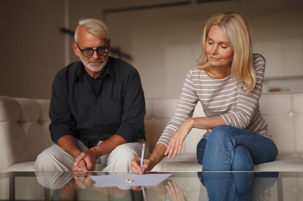 An older woman signs a document on a glass table, while an older man watches attentively. They sit on a cream-colored sofa in a softly lit living room.
