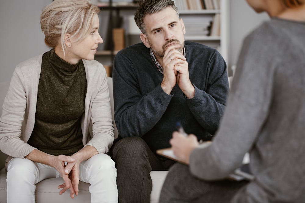 A couple sits attentively, hands clasped, facing a person taking notes. They are in a cozy room with bookshelves, suggesting a counseling or therapy session.