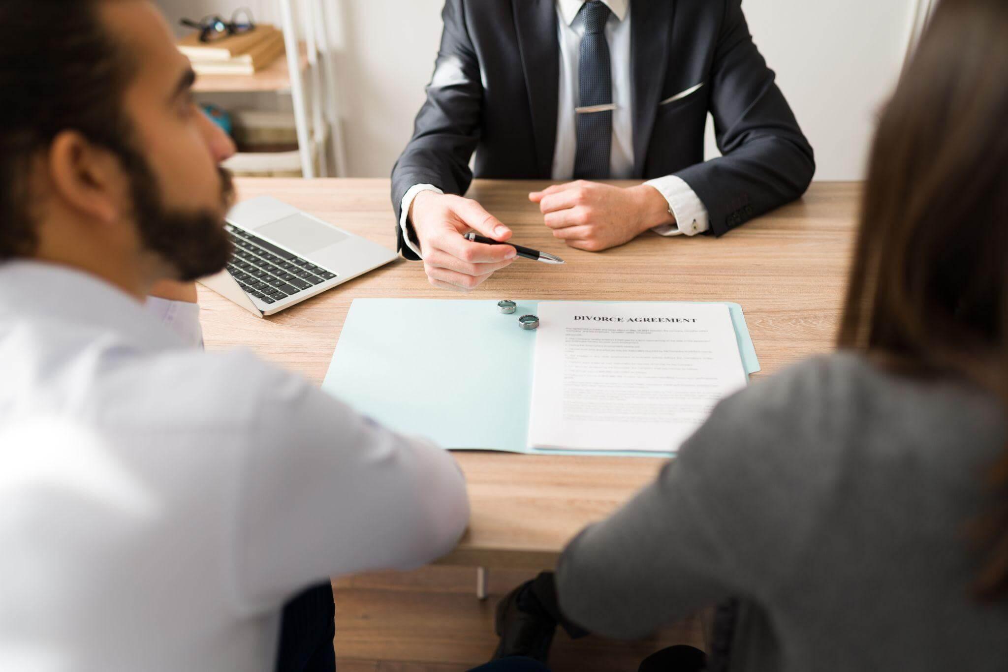 a man in a suit sitting at a table in front of a laptop, a couple is ready to sign a separation document
